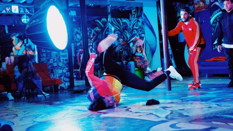 Danseur de breakdance réalisant une figure au sol lors d’un battle hip-hop, entouré de spectateurs dans un lieu décoré de graffiti.