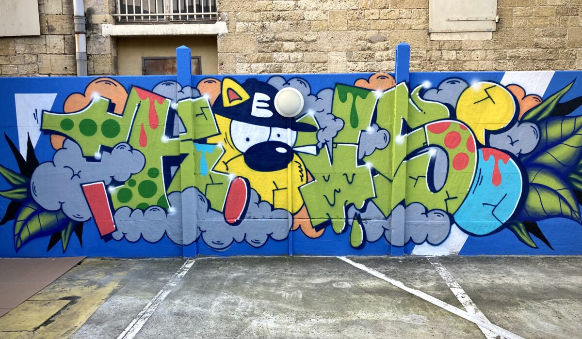 Lettrage graffiti colorée avec personnage cartoon, nuages et feuilles tropicales sur mur bleu à Bordeaux.