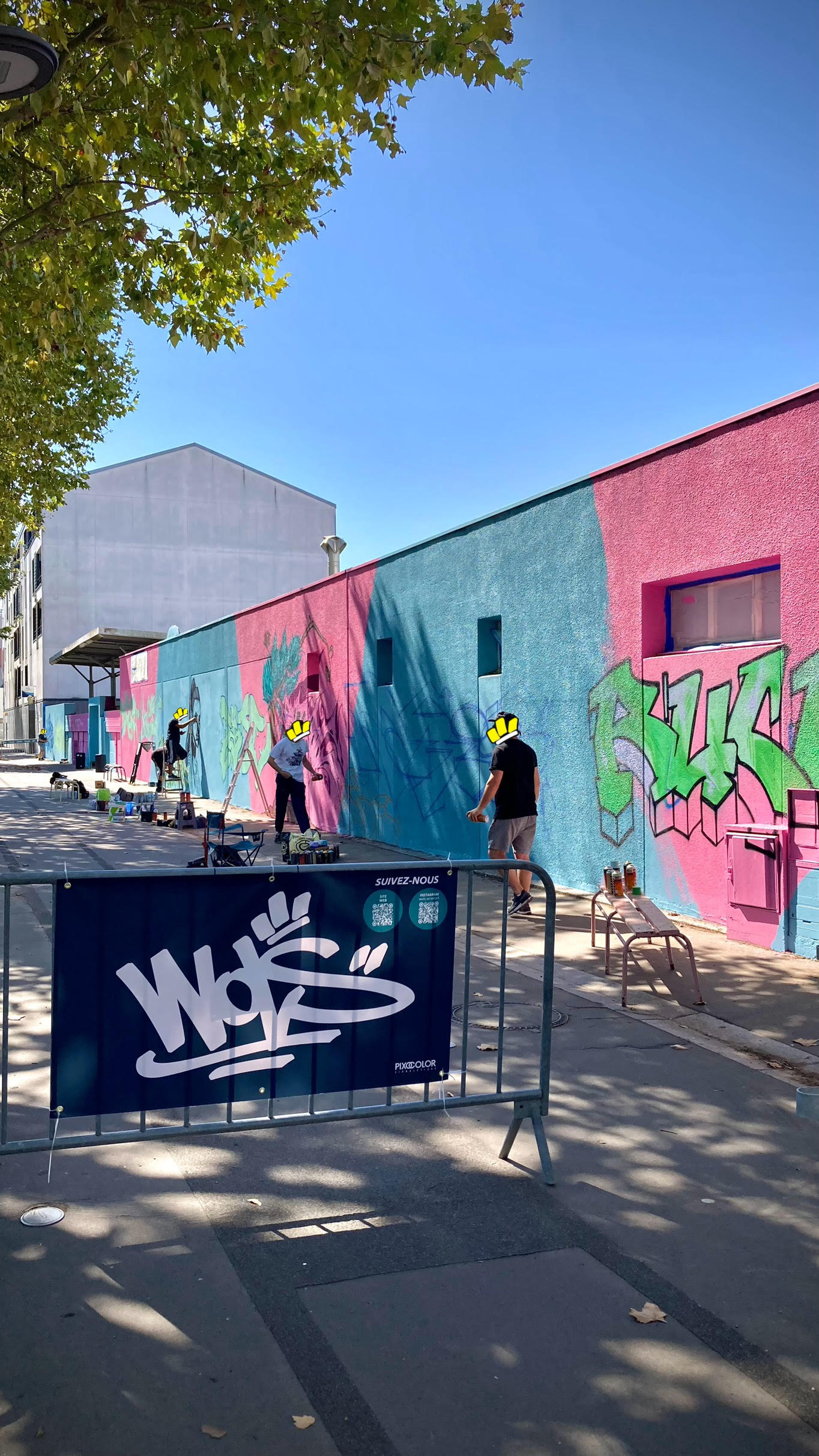 Artistes WOLS peignant une fresque murale colorée sur fond bleu et rose avec matériel de peinture et bombes de graffiti