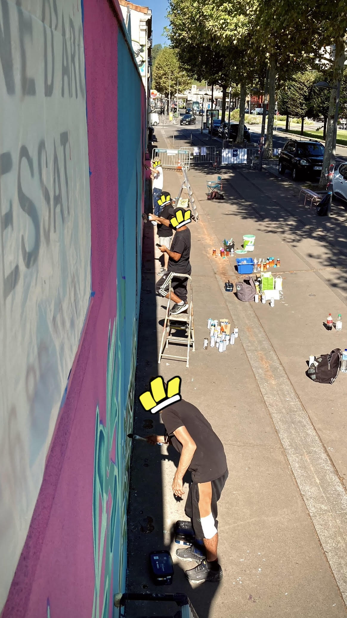 Artistes sur escabeaux réalisant les détails d’une fresque murale avec bombes de peinture et matériel au sol