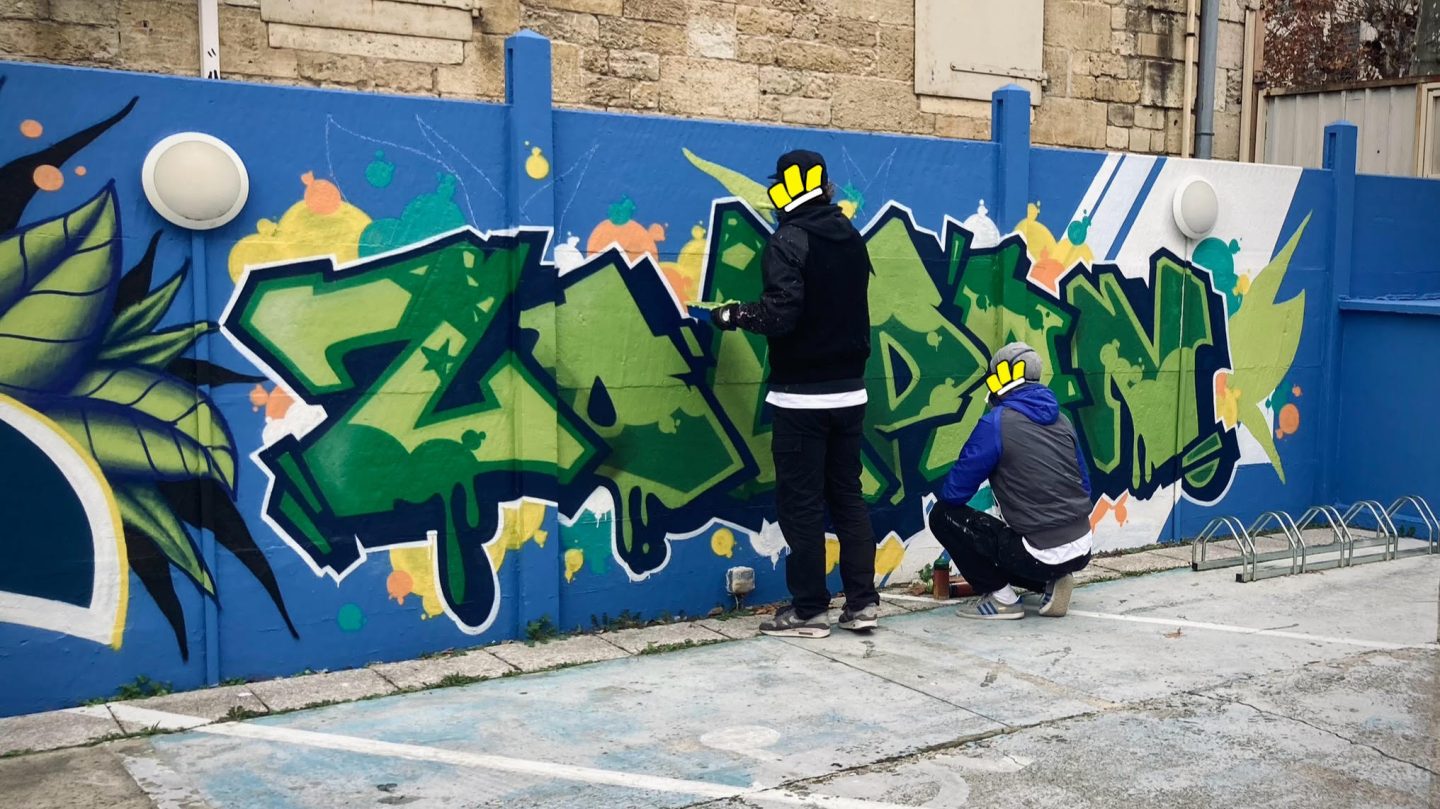 Artistes peignant un lettrage graffiti vert sur mur bleu lors d’un atelier street art public à Bordeaux.