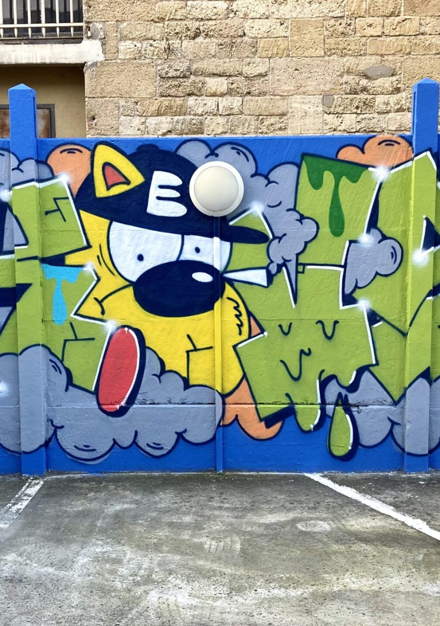 Lettrage graffiti colorée avec personnage cartoon, nuages et feuilles tropicales sur mur bleu à Bordeaux.