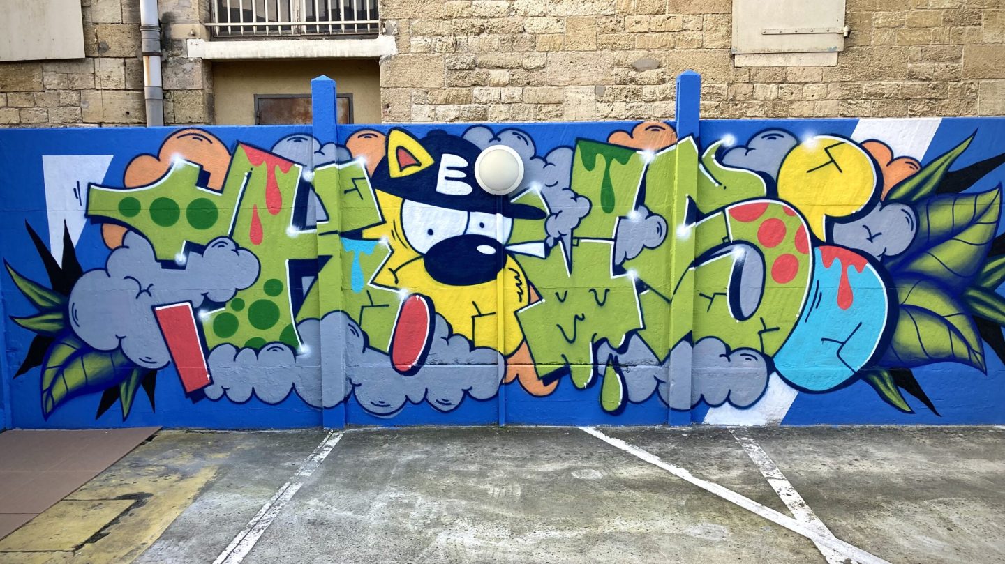 Lettrage graffiti colorée avec personnage cartoon, nuages et feuilles tropicales sur mur bleu à Bordeaux.