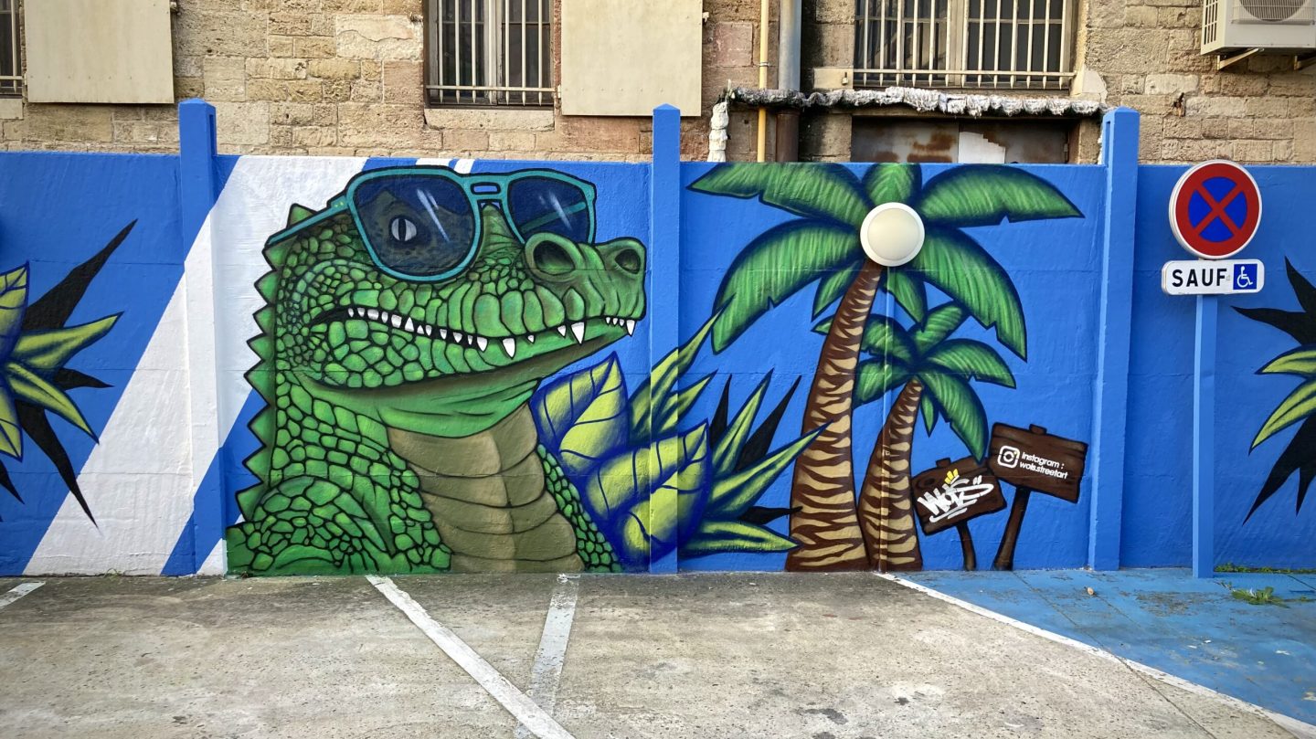 Crocodile graffiti avec lunettes de soleil et palmiers peint sur le mur bleu du parking Zolpan Bordeaux.