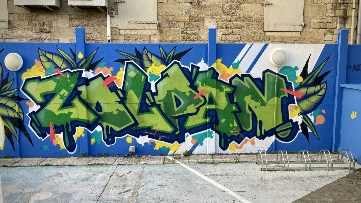 Lettrage graffiti vert Zolpan sur mur bleu avec feuilles tropicales, éclats colorés et style street art contemporain.