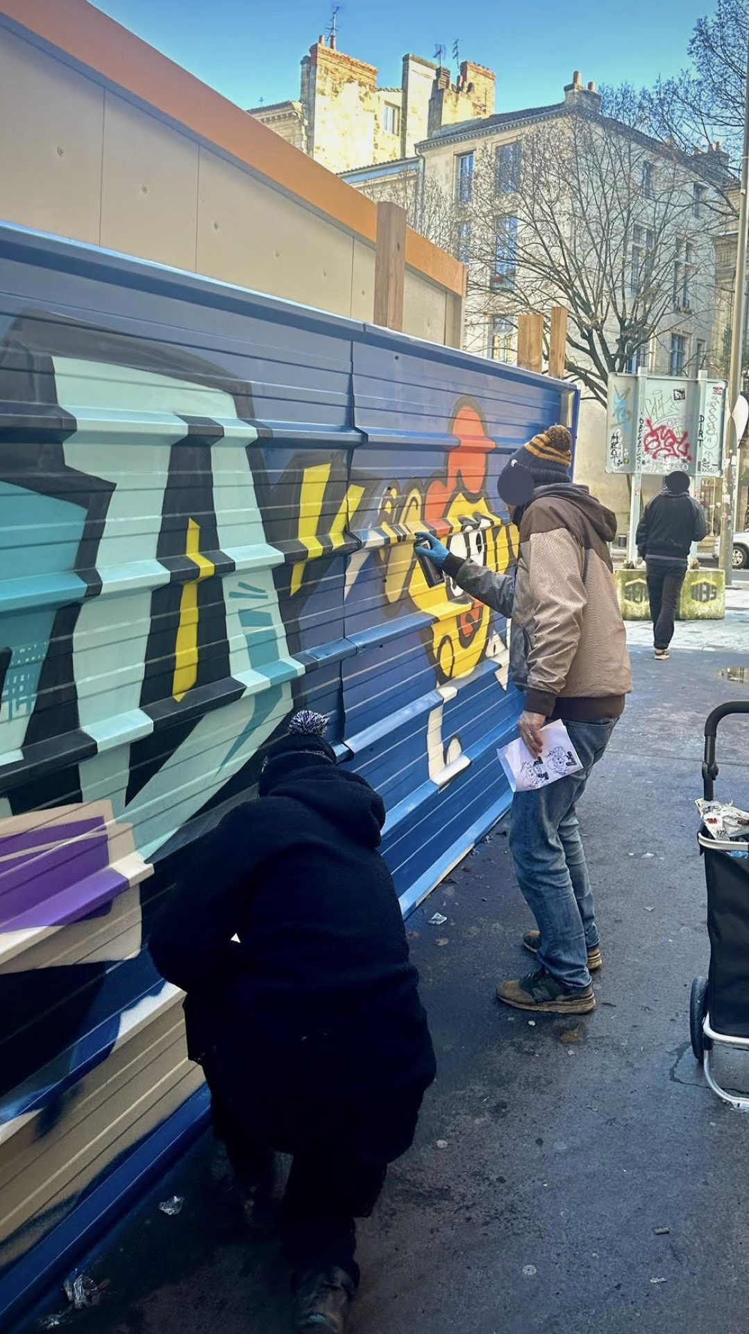 Artistes graffiti MEL20 et Monsieur Poulet réalisant une fresque street art collaborative sur une palissade métallique de chantier à Bordeaux en décembre 2024 pour habiller l’espace urbain et prévenir les tags sauvages.