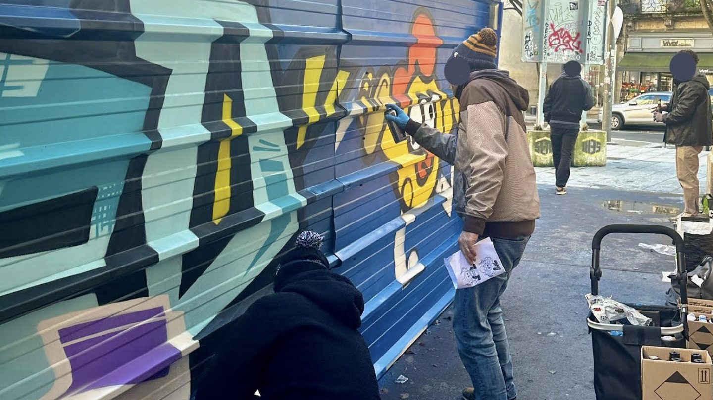 Artistes graffiti MEL20 et Monsieur Poulet réalisant une fresque street art collaborative sur une palissade métallique de chantier à Bordeaux en décembre 2024 pour habiller l’espace urbain et prévenir les tags sauvages.