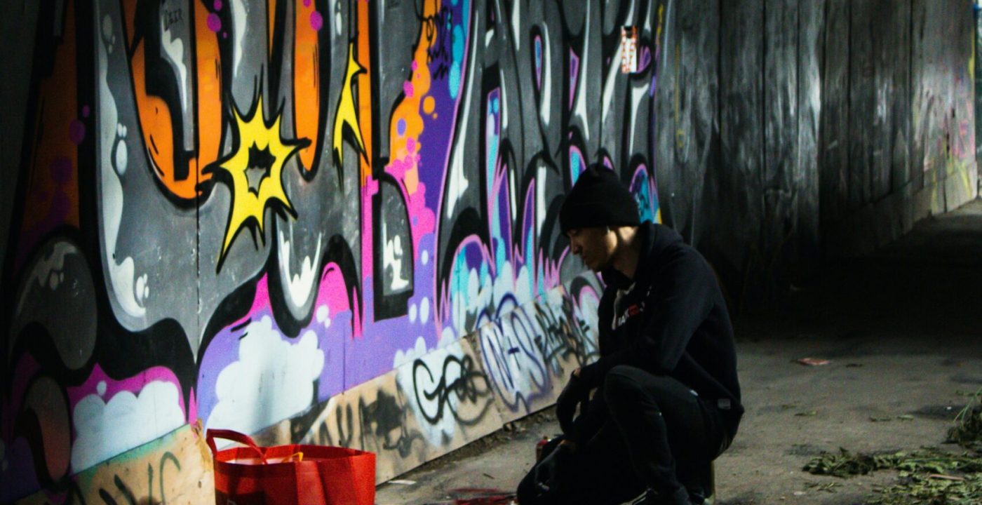 Artiste graffiti réalisant une fresque street art colorée dans un lieu urbain industriel avec bombes de peinture aérosol.
