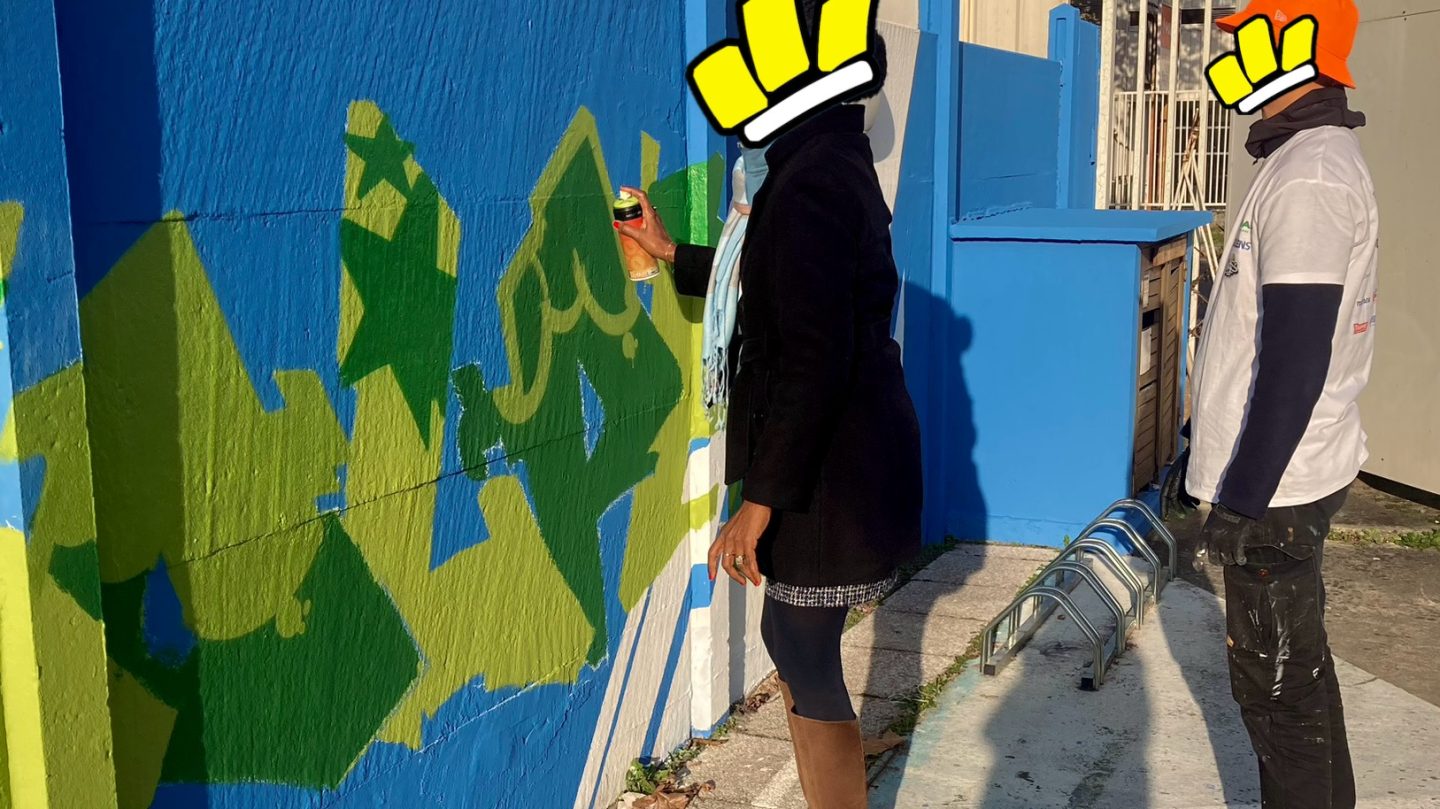 Participante initiée au graffiti peignant un lettrage vert sur mur bleu lors d’un atelier street art à Bordeaux.