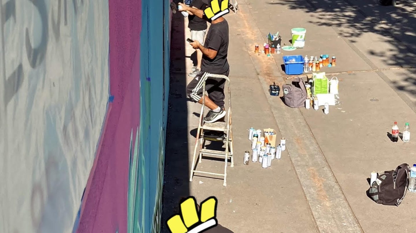 Artistes sur escabeaux réalisant les détails d’une fresque murale avec bombes de peinture et matériel au sol