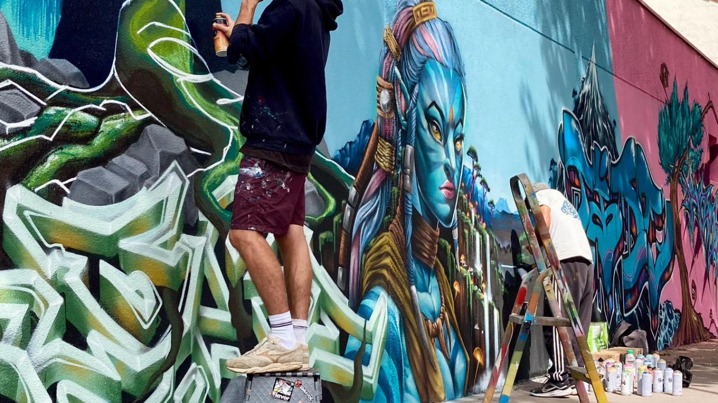 Artistes réalisant une fresque Avatar avec portrait et graffiti sur un mur extérieur