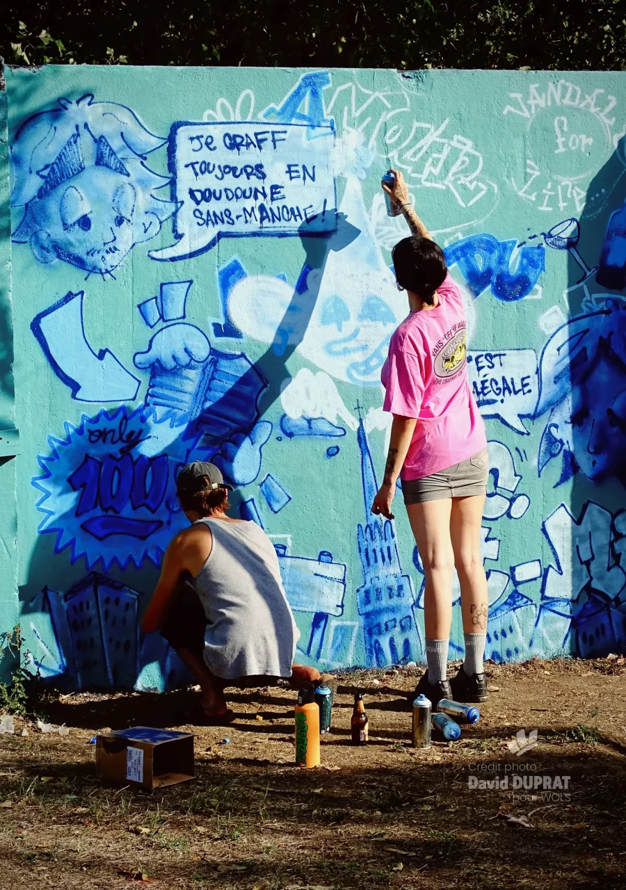 Deux participants peignant à la bombe un mur couvert de dessins et de graffiti lors du WOLS Open Spray #2.