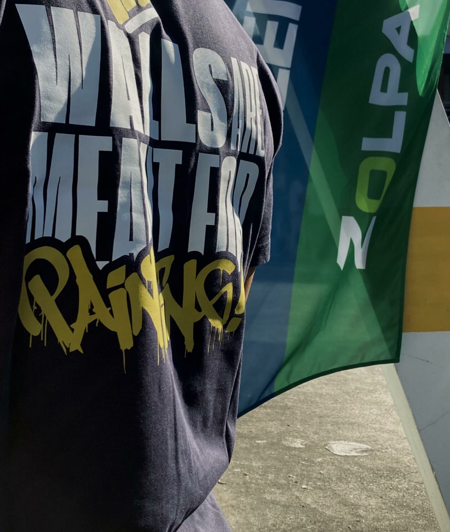 Personne portant un tee-shirt WOLS Streetwear avec le slogan “Walls Are Meant For Painting” devant une oriflamme Tollens-Zolpan.