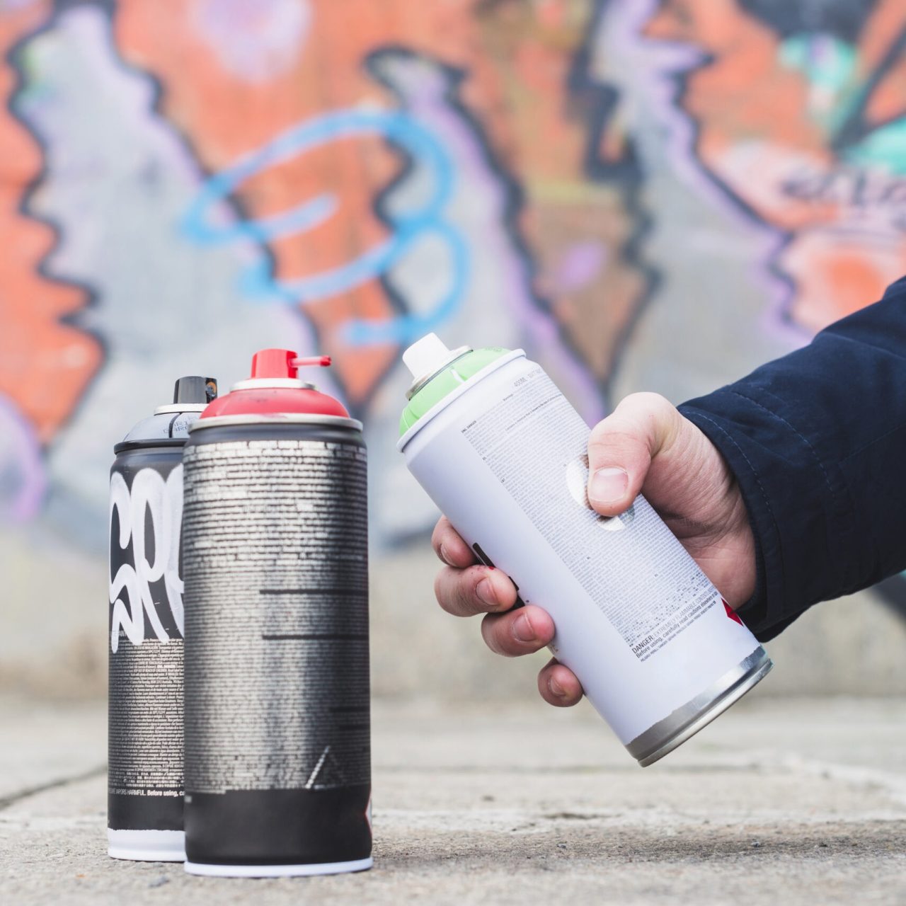 Main tenant une bombe de peinture aérosol devant un mur graffiti coloré avec plusieurs sprays posés au sol.