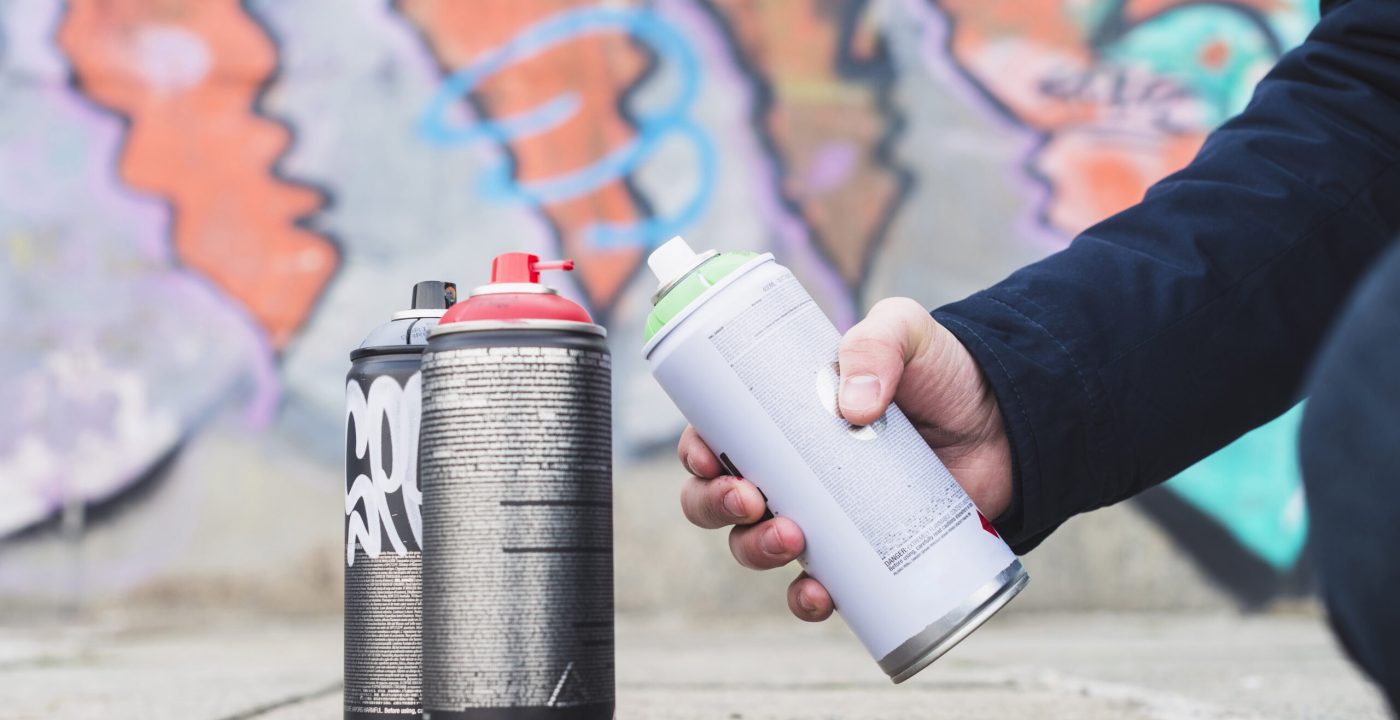 Main tenant une bombe de peinture aérosol devant un mur graffiti coloré avec plusieurs sprays posés au sol.